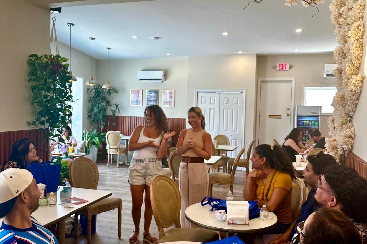 People gathered in a cafe, two stand talking, others seated at round tables.